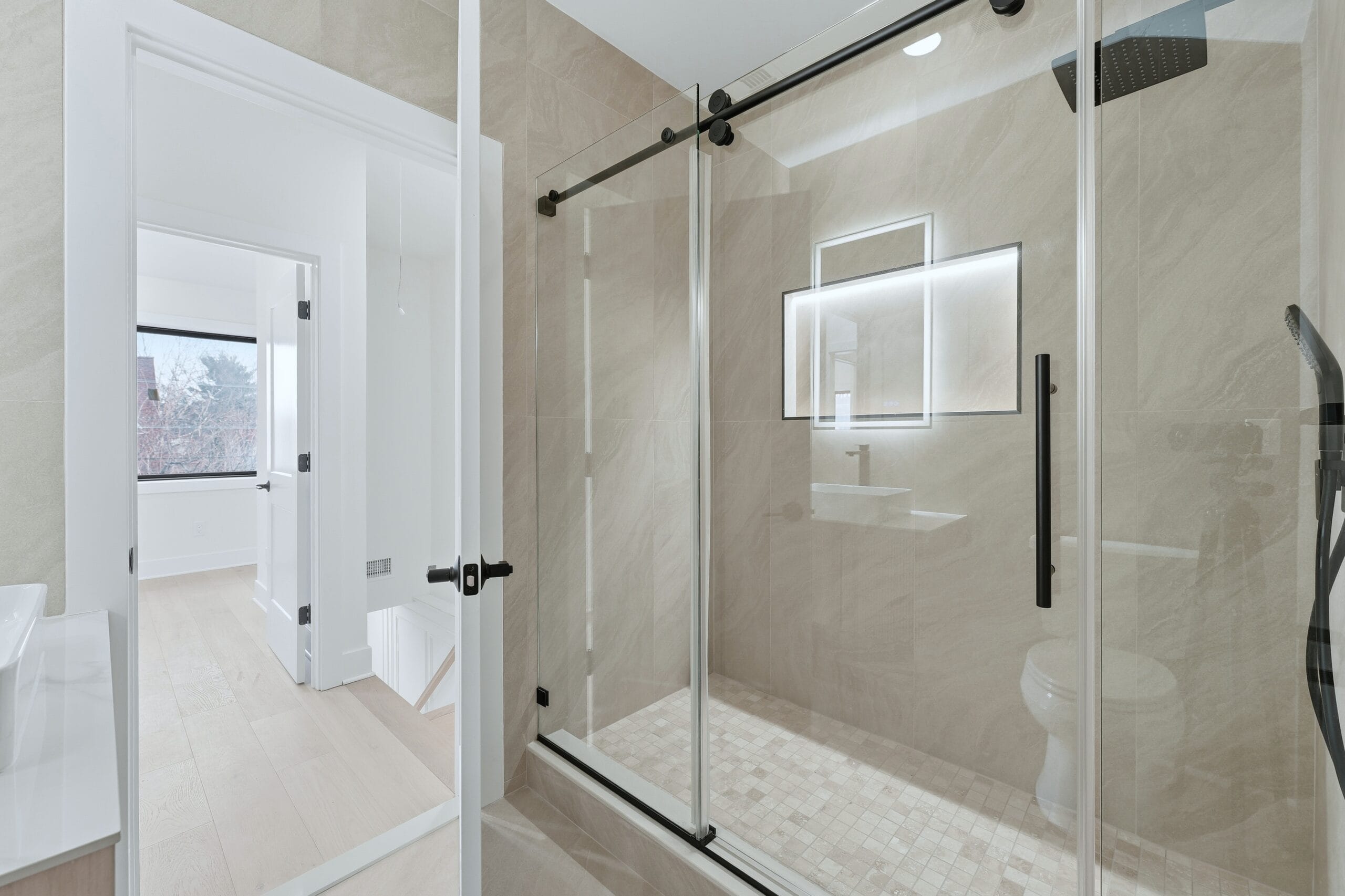 Marble-tiled shower with a sliding glass door, backlit niche mirror, and matte-black hardware